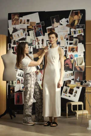 Fashion designer fits a white dress on a model in a studio setting, mood board with images in the background, natural light.