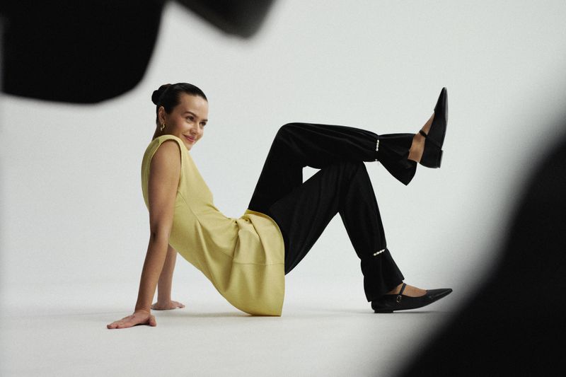 A stylish young woman in a light yellow sleeveless top and black wide-legged pants poses in a photo studio against a bright background.