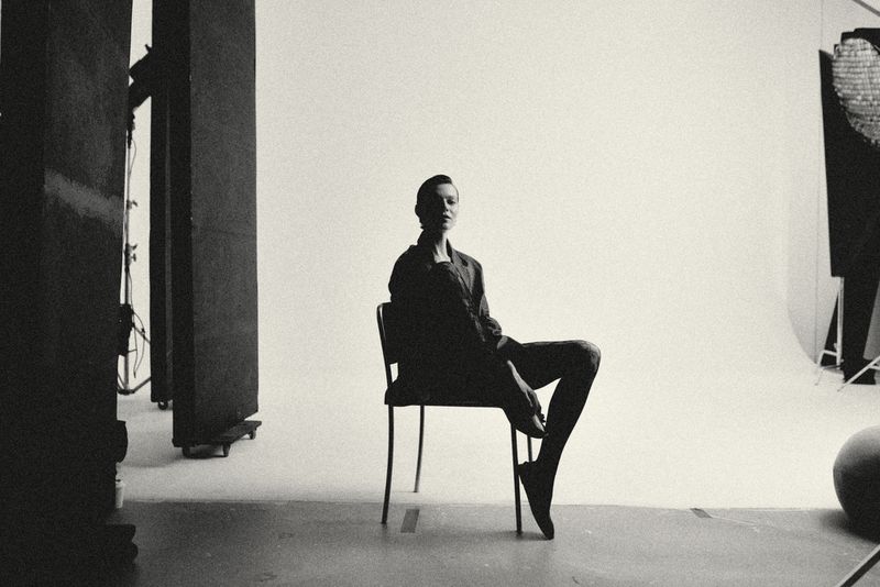 Monochrome studio shot of a female model in a blazer sitting on a chair, highlighting fashion and minimalist aesthetics in a clean environment.