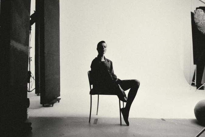 Monochrome studio shot of a female model in a blazer sitting on a chair, highlighting fashion and minimalist aesthetics in a clean environment.