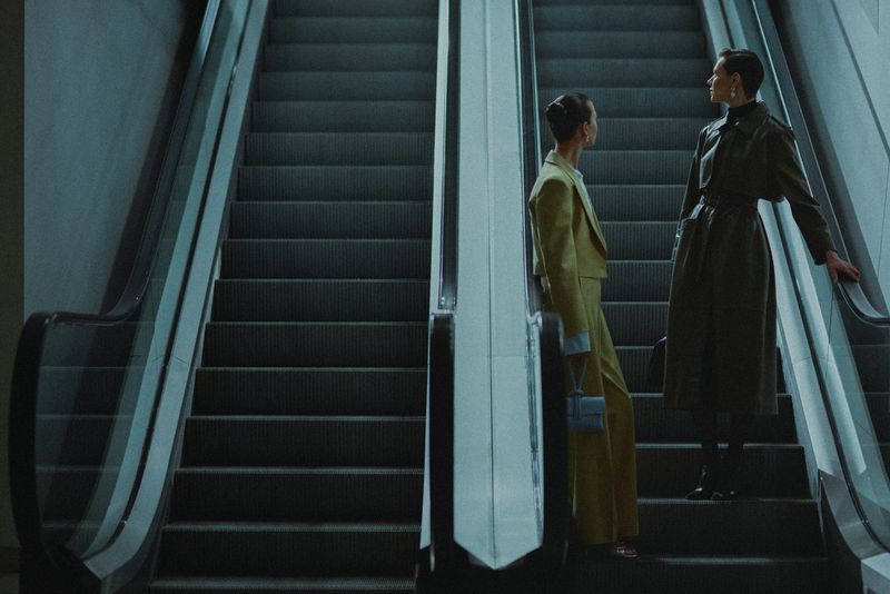 Two women in fashionable attire ascend an escalator in a modern, minimalist setting, bathed in soft, diffused lighting.