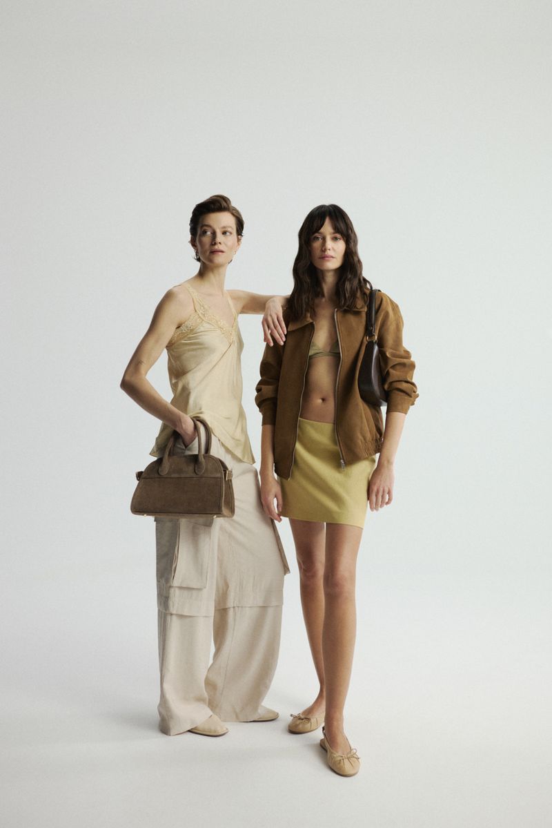 Two women pose in a studio, one in a tank top and cargo pants holding a purse, the other in a jacket and skirt with a purse.
