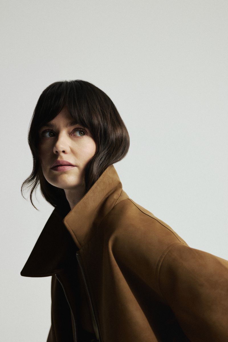 Adult woman in a brown coat looks up with a slight smile against a neutral background in a studio shot with soft, diffused lighting.