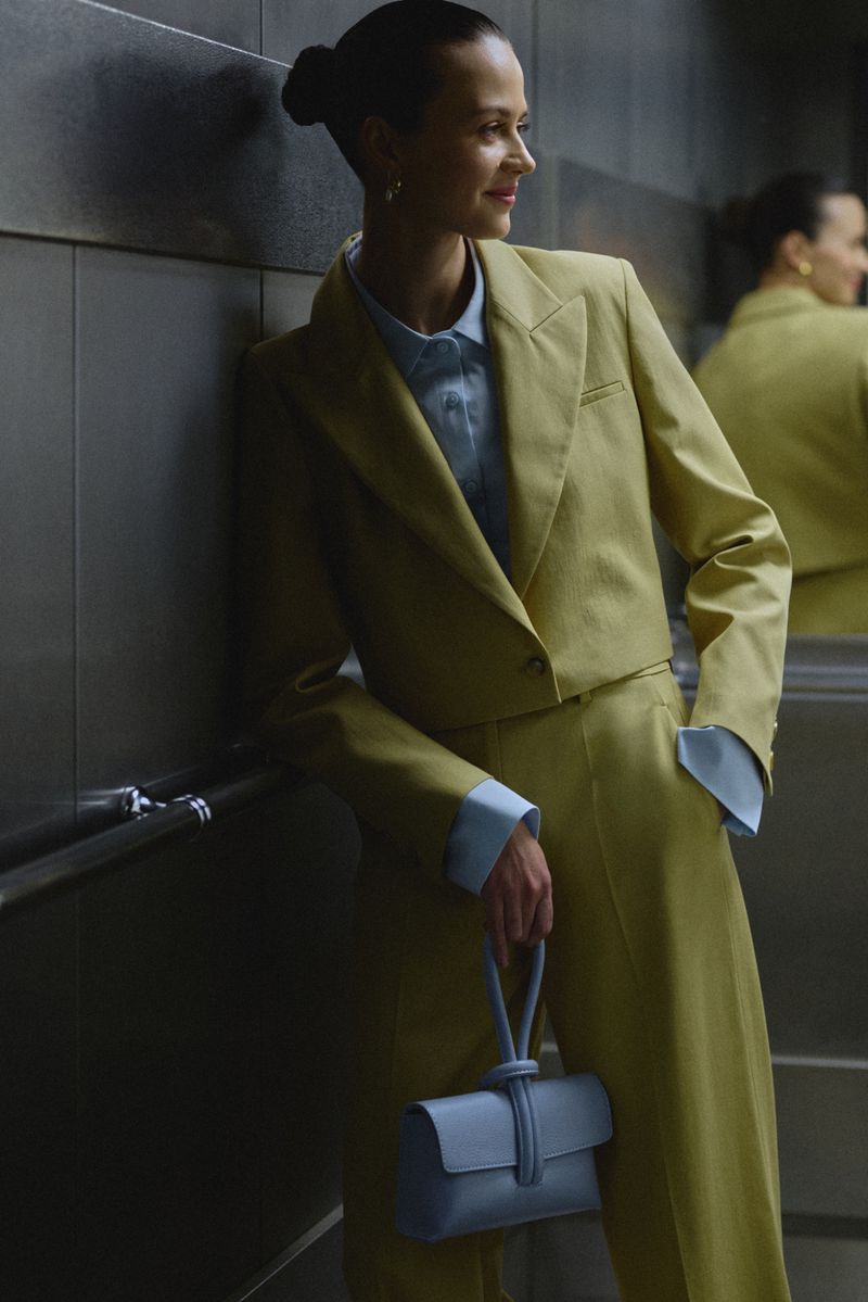 Woman in yellow cropped suit poses indoors with a blue handbag, looking away. Her hair is pulled back and she is wearing a light blue shirt.
