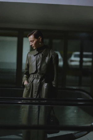 A stylish woman in a belted olive trench coat stands on an escalator in an indoor environment, looking down with a handbag.