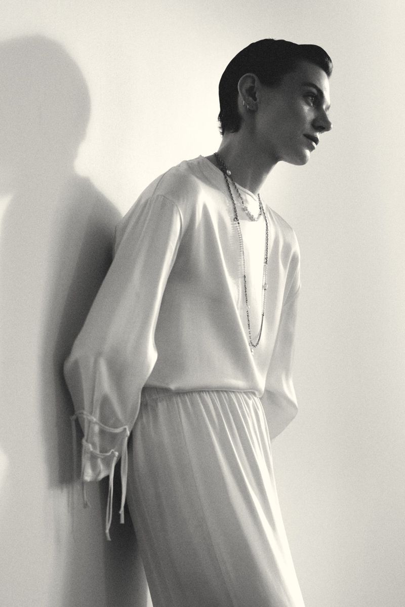 Model Marta Dyks in a draped white silk top with layered necklaces leaning against a wall in a black and white fashion portrait.