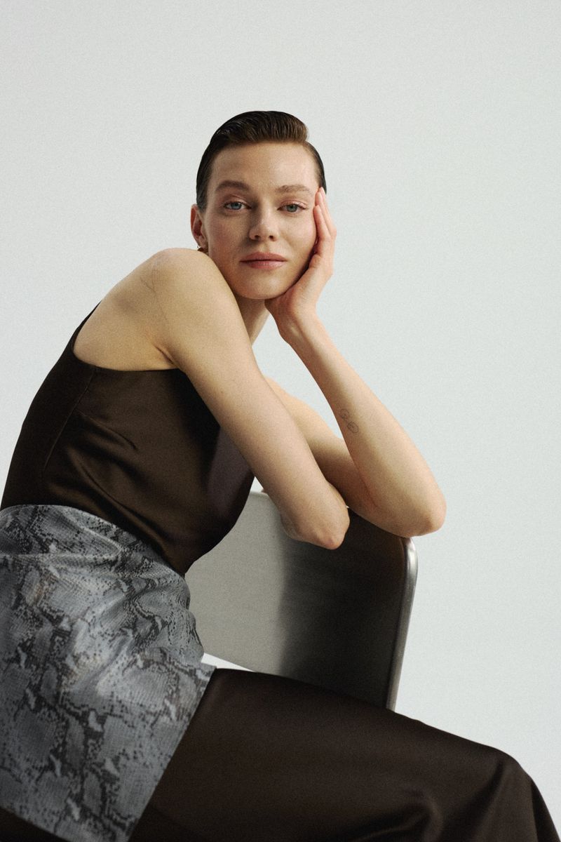 Adult woman with short brown hair poses seated on a chair in a photo studio, wearing a brown tank top and patterned skirt against a white backdrop.