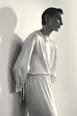 Model Marta Dyks in a draped white silk top with layered necklaces leaning against a wall in a black and white fashion portrait.