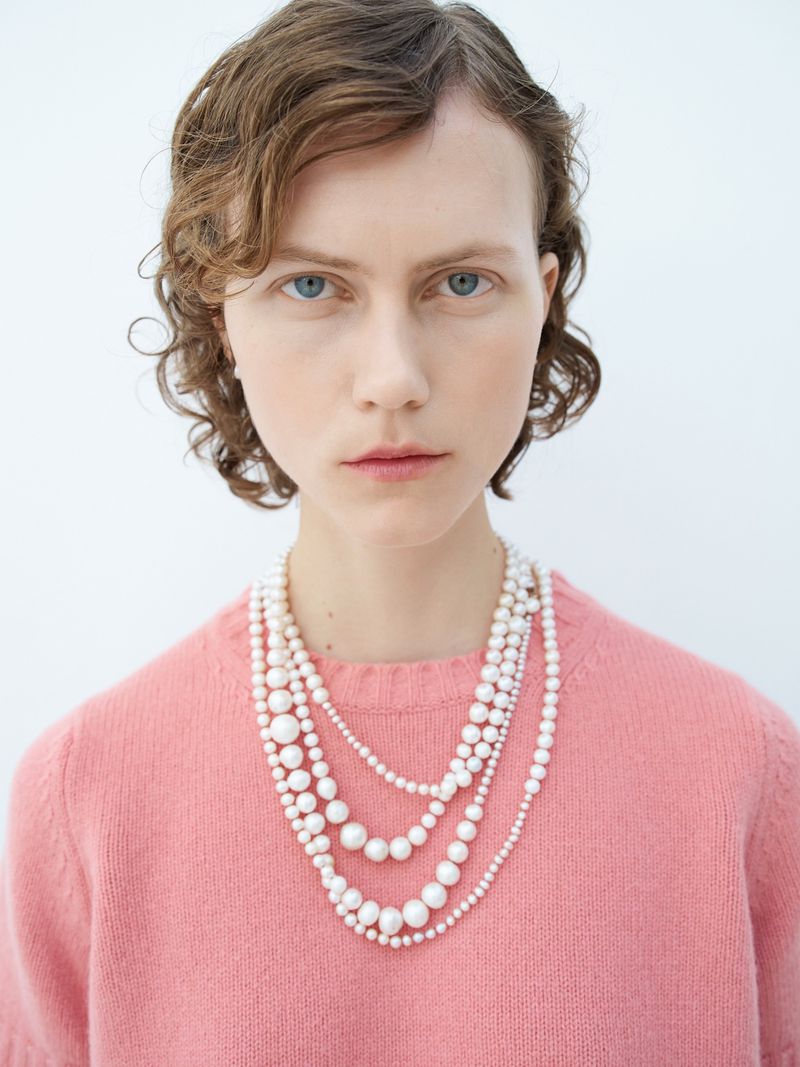 Close-up of a young woman with curly brown hair and blue eyes, wearing a pink sweater and layered pearl necklaces against white backdrop.
