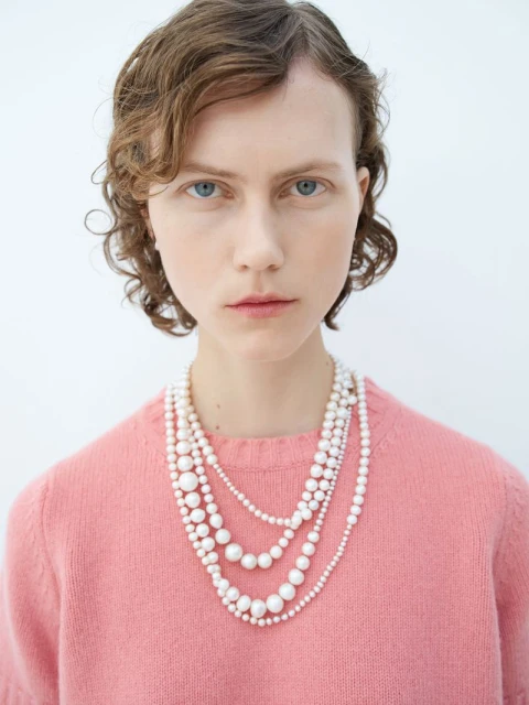 Close-up of a young woman with curly brown hair and blue eyes, wearing a pink sweater and layered pearl necklaces against white backdrop.