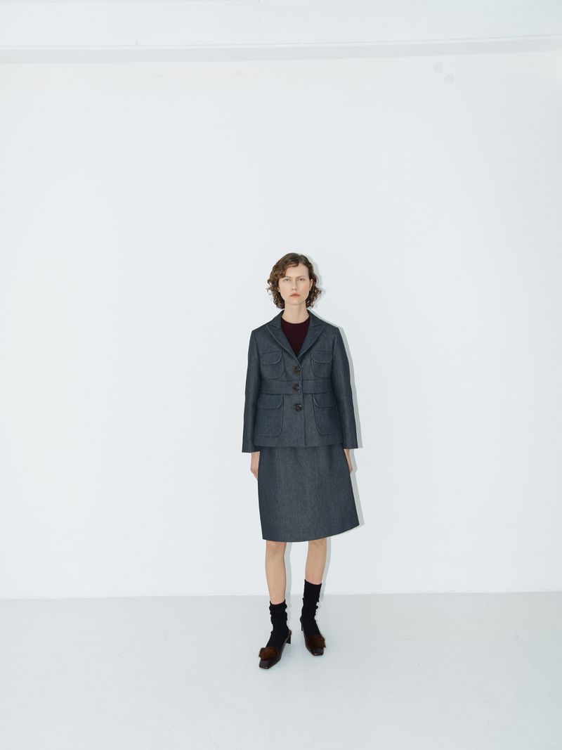 Woman stands in a photo studio wearing a gray skirt suit with a burgundy top, black socks, and brown shoes against a white backdrop.
