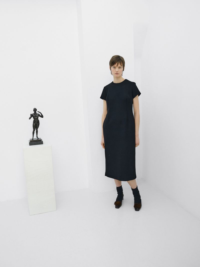 Indoor shot of a female adult in a navy dress and statue on a white pedestal in a white corner space, featuring neutral expression.