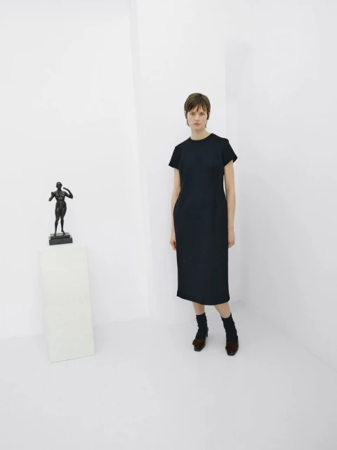 Indoor shot of a female adult in a navy dress and statue on a white pedestal in a white corner space, featuring neutral expression.