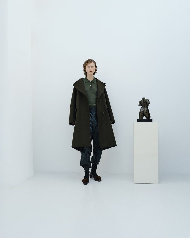 Woman in a long coat standing next to a small statue in a minimalist studio setting with white walls and floors and neutral light.
