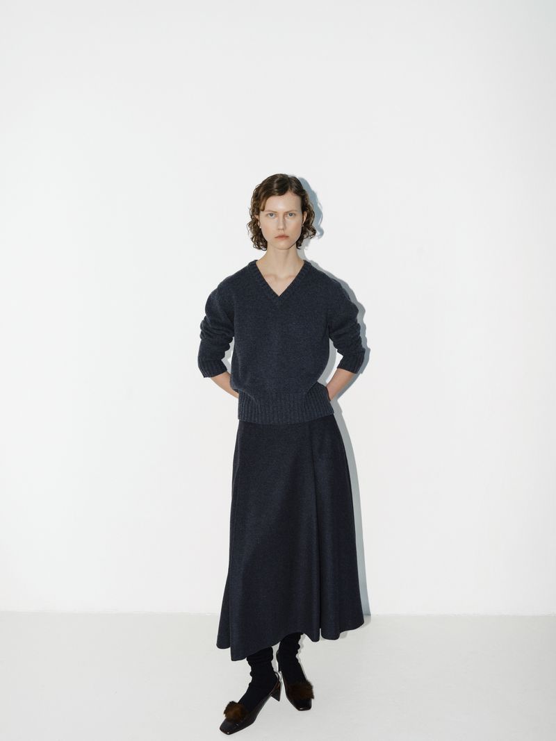 A woman poses in a fashion studio wearing a navy knit sweater and matching long skirt, complemented by dark socks and distinctive heeled shoes.