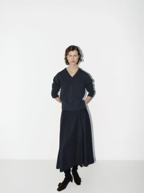 A woman poses in a fashion studio wearing a navy knit sweater and matching long skirt, complemented by dark socks and distinctive heeled shoes.