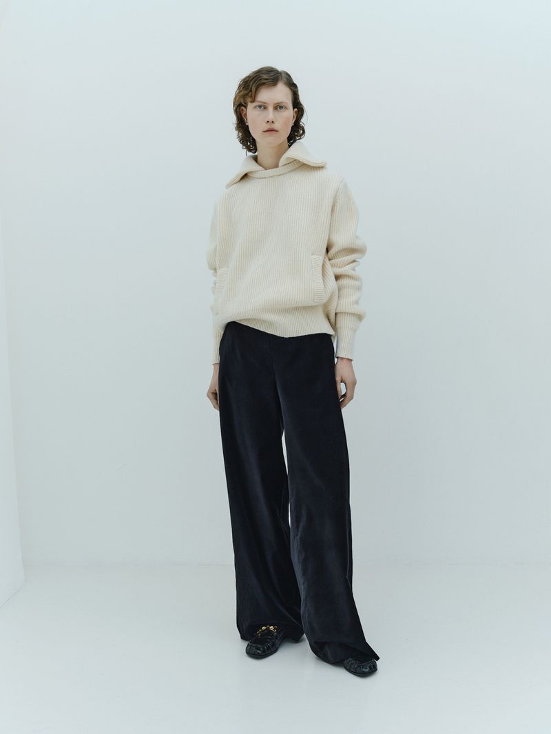 Full-length studio shot of a woman in a cream-colored sweater and wide-legged black pants, posing against a light grey seamless backdrop.
