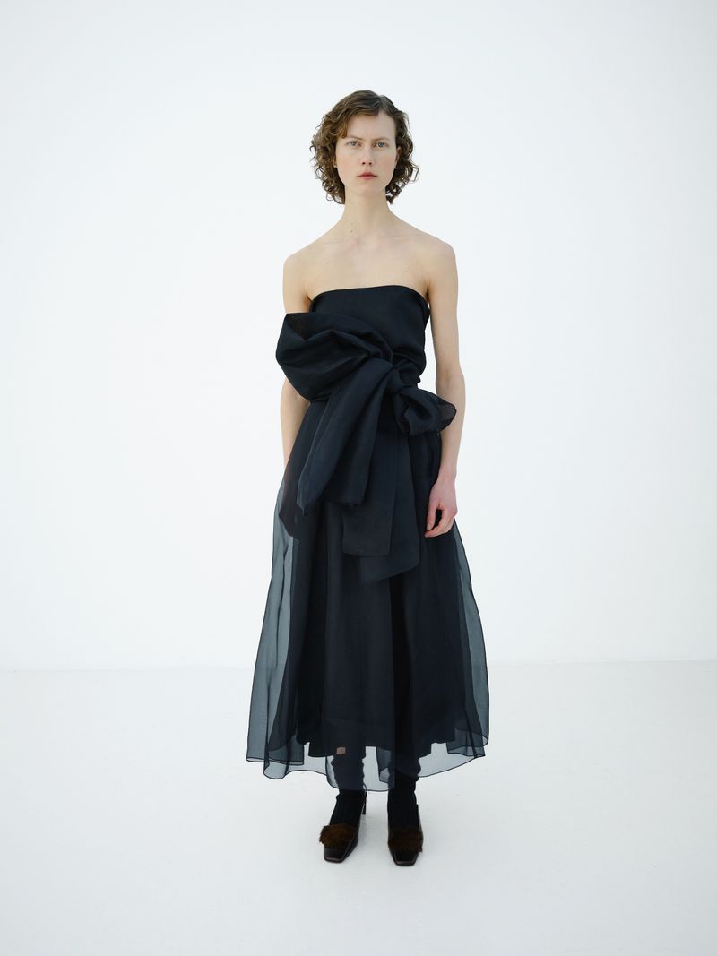 An adult woman is posing in a strapless black dress with a bow-like feature in a white photo studio with professional lighting.