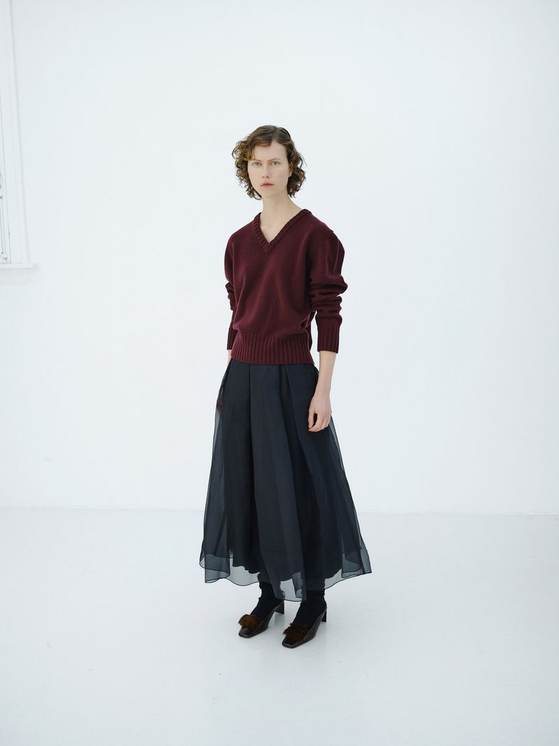 A woman in a burgundy sweater and black culottes stands in a white room, fashion editorial, studio lighting, and understated elegance.