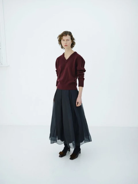 A woman in a burgundy sweater and black culottes stands in a white room, fashion editorial, studio lighting, and understated elegance.