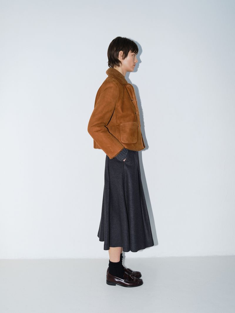 Woman poses in brown suede jacket and grey skirt against a white background in a studio shot, fashion editorial.