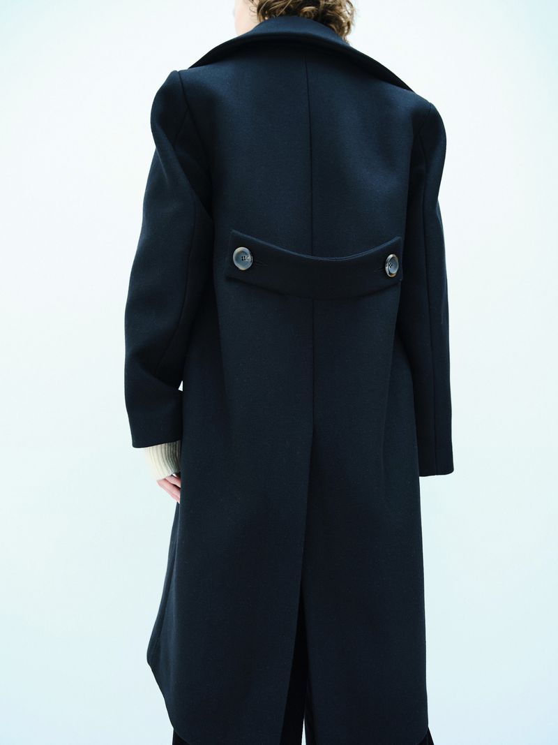 Model showcases a dark, long-sleeved, buttoned overcoat against a clean white backdrop in a fashion-forward studio shot.