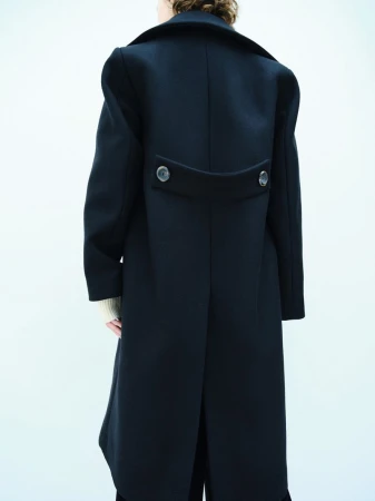 Model showcases a dark, long-sleeved, buttoned overcoat against a clean white backdrop in a fashion-forward studio shot.