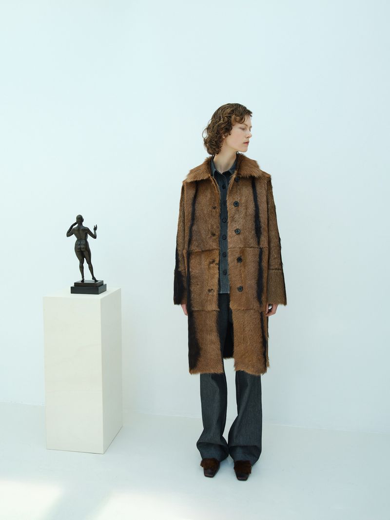 Woman in a brown faux fur coat posing in a minimal white photo studio next to a sculpture on a pedestal, with a clean aesthetic.