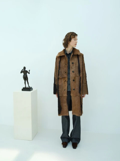 Woman in a brown faux fur coat posing in a minimal white photo studio next to a sculpture on a pedestal, with a clean aesthetic.