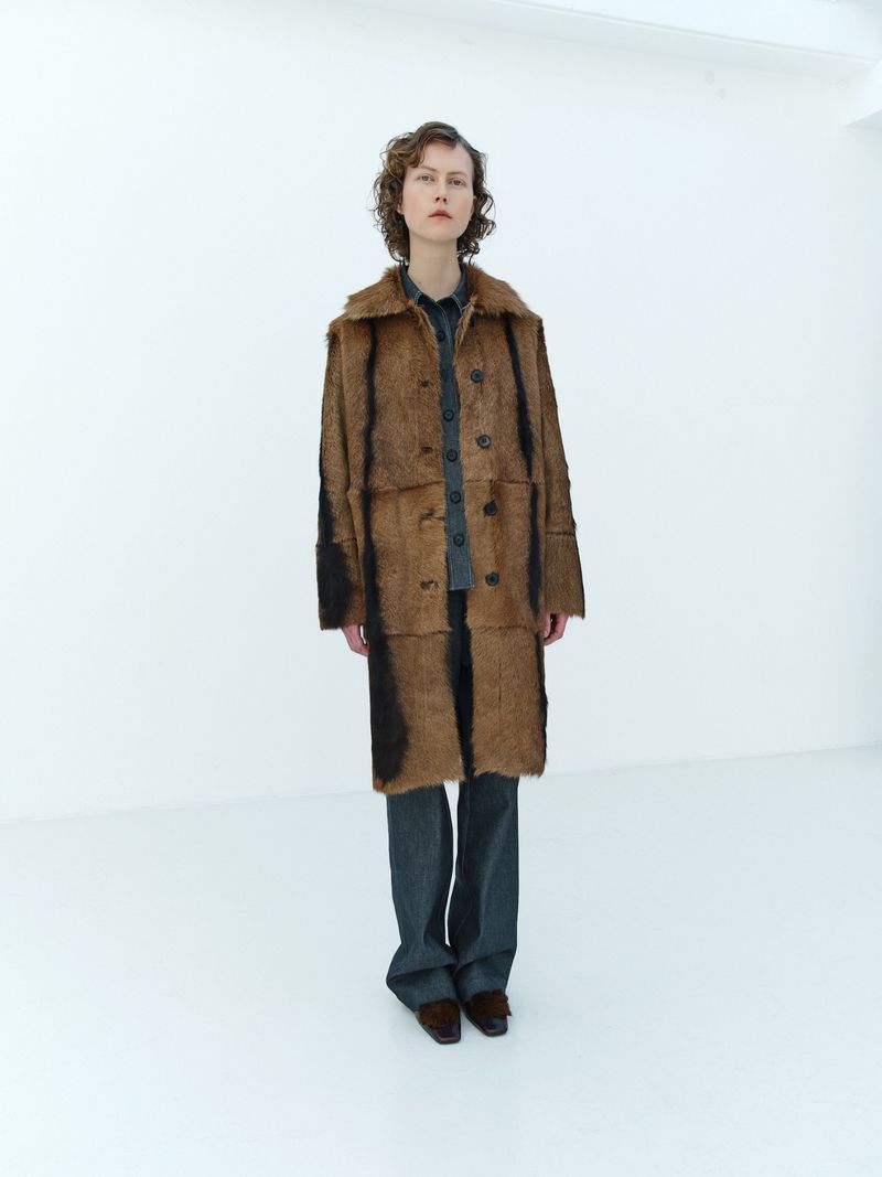 Full shot of a woman in a brown patterned coat, gray pants, and brown shoes posing in a white photo studio with minimalist aesthetic.