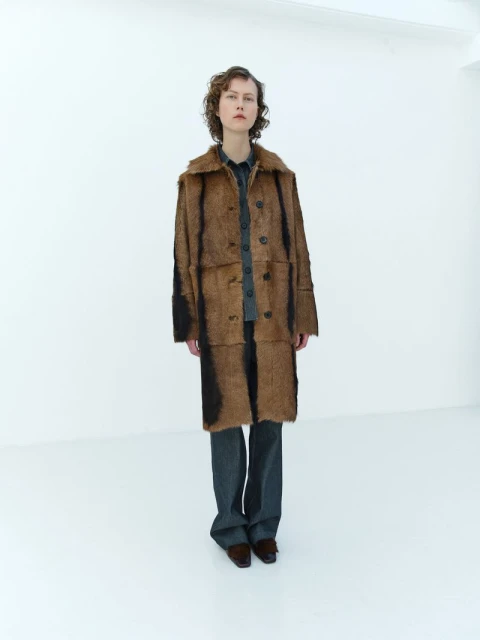 Full shot of a woman in a brown patterned coat, gray pants, and brown shoes posing in a white photo studio with minimalist aesthetic.