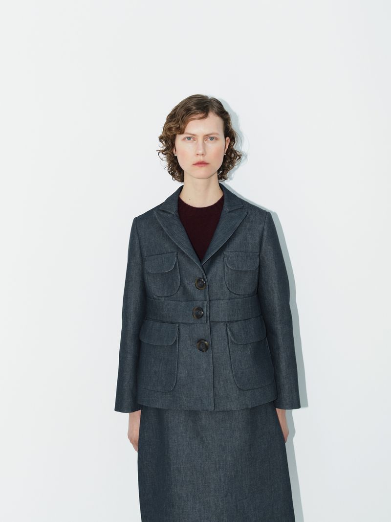 A woman in a dark denim skirt suit with patch pockets stands against a white background in a fashion studio shot with minimal styling.