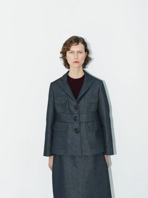 A woman in a dark denim skirt suit with patch pockets stands against a white background in a fashion studio shot with minimal styling.