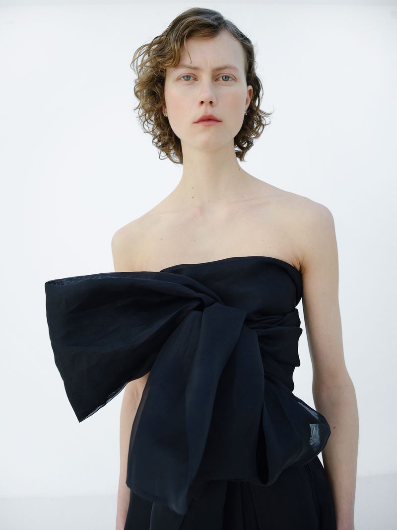 A light-skinned woman with curly short hair wearing a strapless black top with a large bow detail, posing against a plain white background.
