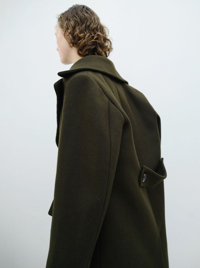 Model seen from behind wearing an olive green tailored wool coat against a pale grey seamless studio backdrop.