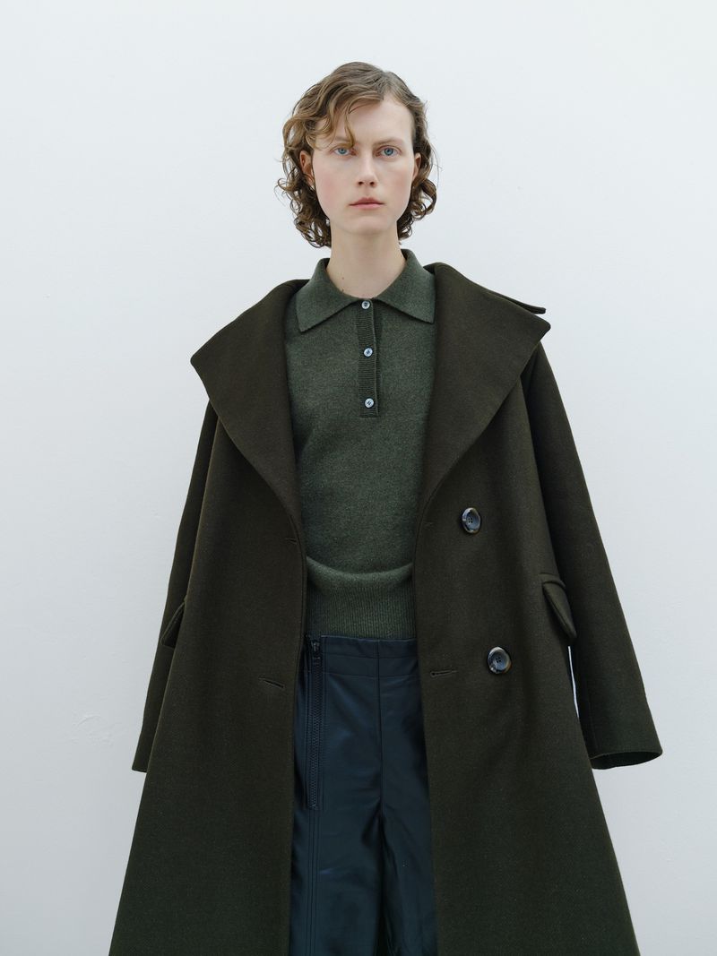 A woman in a green collared shirt and a large dark green coat poses in front of a white wall in a fashion photography shot.
