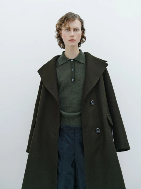 A woman in a green collared shirt and a large dark green coat poses in front of a white wall in a fashion photography shot.