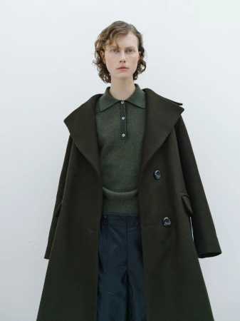 A woman in a green collared shirt and a large dark green coat poses in front of a white wall in a fashion photography shot.