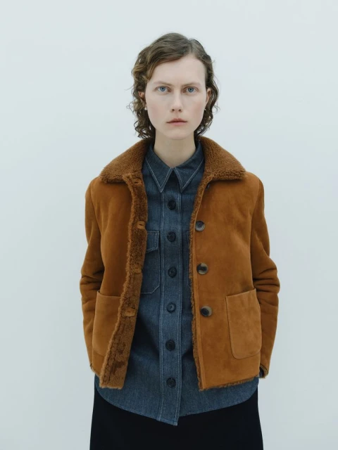 A woman with curly hair poses in a studio wearing a brown shearling jacket over a denim shirt and a black skirt on a white background.