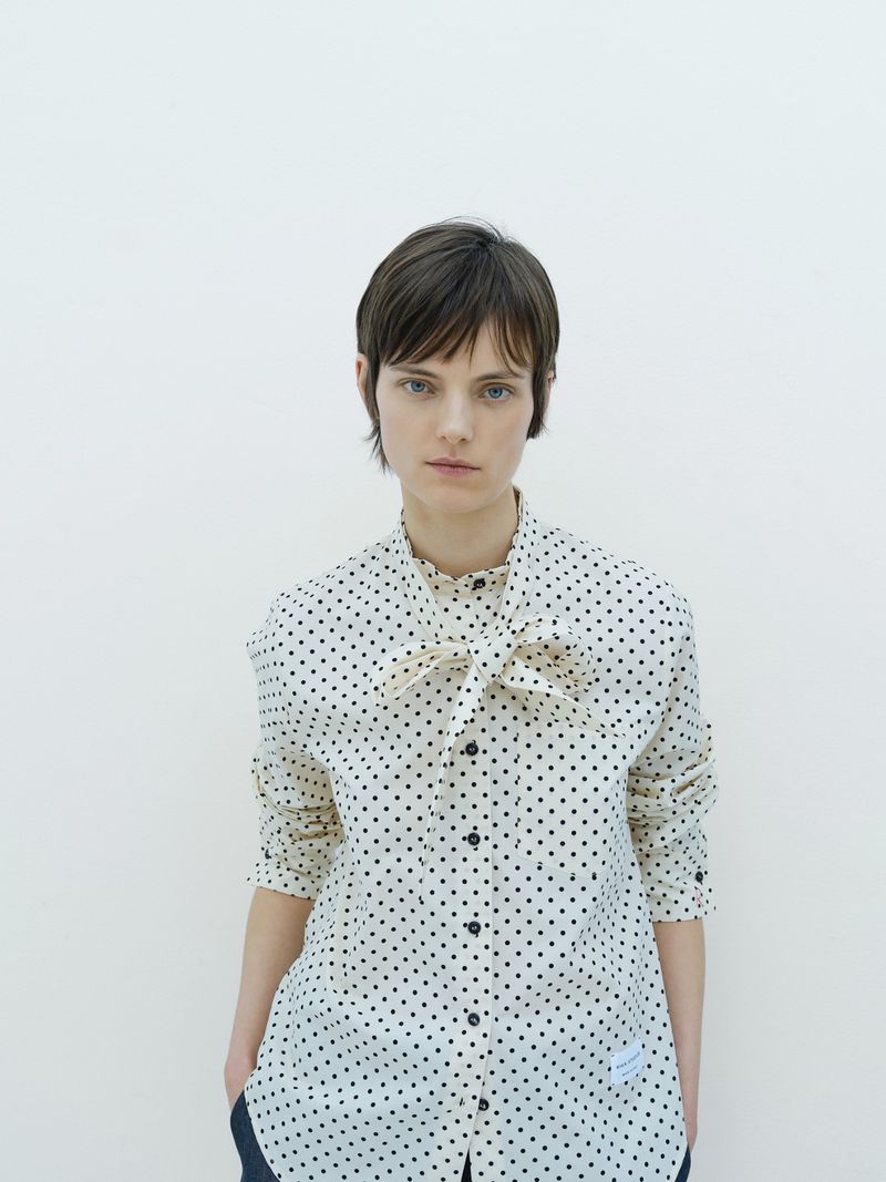Woman in a polka dot shirt with a bow tie poses against a white wall, fashion editorial, eye-level angle, studio lighting, portrait orientation.