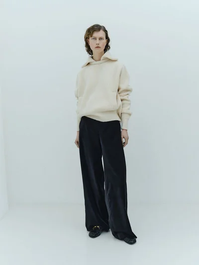 Model wearing a cream ribbed sweater and dark trousers standing against a seamless white studio background.