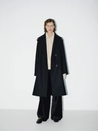 Model wearing a long black coat, cream sweater, and black trousers against a plain white studio background