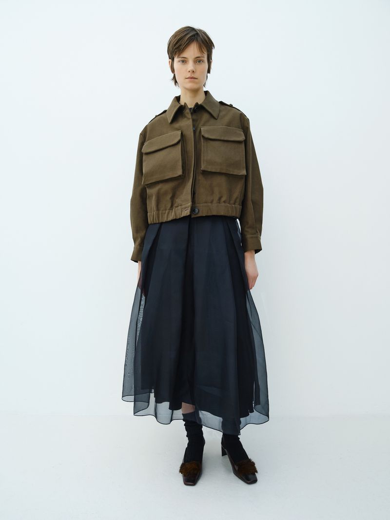 Full-length portrait of a model in a cropped brown utility jacket and sheer layered black skirt standing against a white studio backdrop.