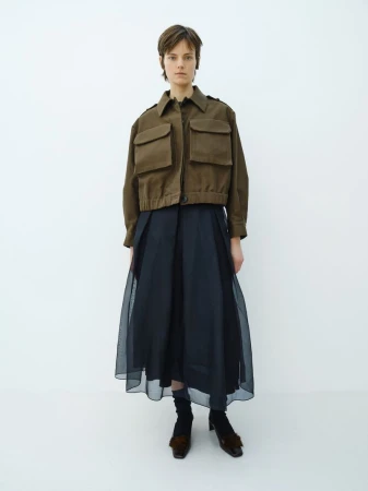 Full-length portrait of a model in a cropped brown utility jacket and sheer layered black skirt standing against a white studio backdrop.