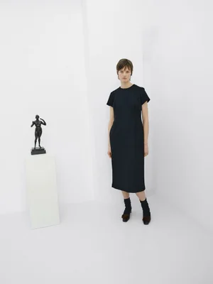 Model in a black short-sleeve midi dress standing beside a small bronze sculpture on a white pedestal in a bright white studio.