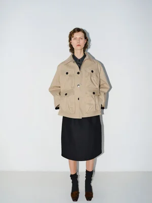 Model standing against a white backdrop wearing a camel utility jacket and a black midi skirt with black socks and shoes.