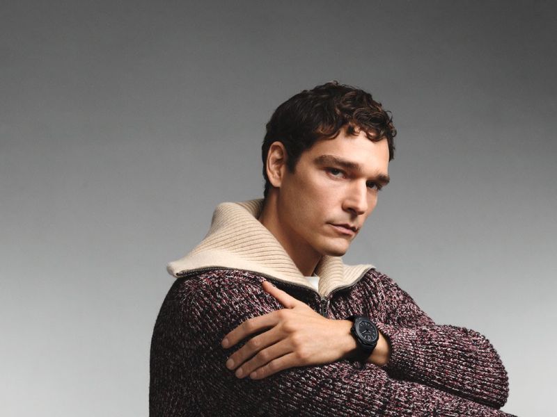 Close-up of a dark-haired adult male model wearing a knit zip-up sweater and a black watch, posing in a photo studio setting.