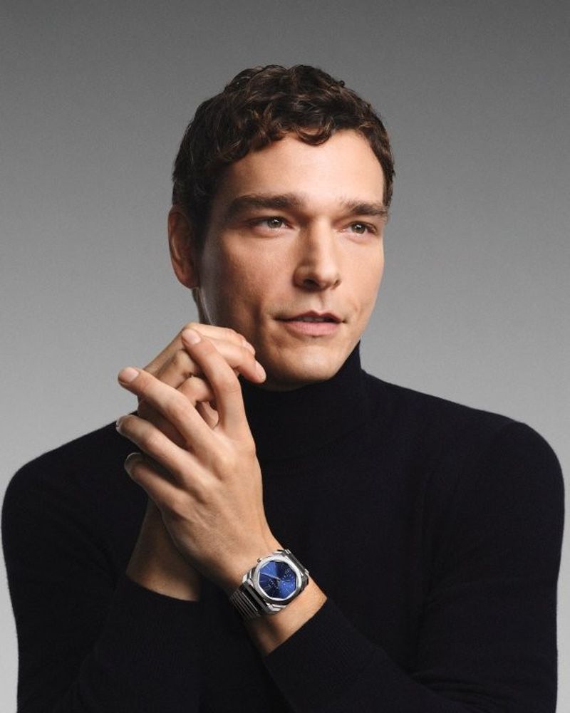 Close-up shot of a man with curly brown hair in a black turtleneck, wearing a silver watch with a blue face, against a gray backdrop.
