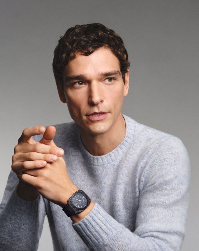 A brunette adult male is posing in a light grey sweater and wearing a watch on his wrist, photographed in a photo studio with soft lighting.