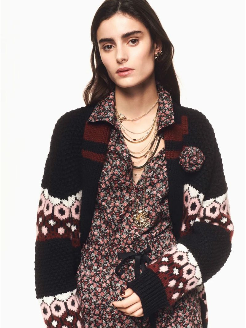 A woman in a floral dress and black cardigan with a rose brooch poses in a studio with layered necklaces for a fashion editorial.
